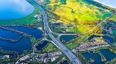 A road and roundabout seen from above, cutting through sunlit green fields and flooded plains